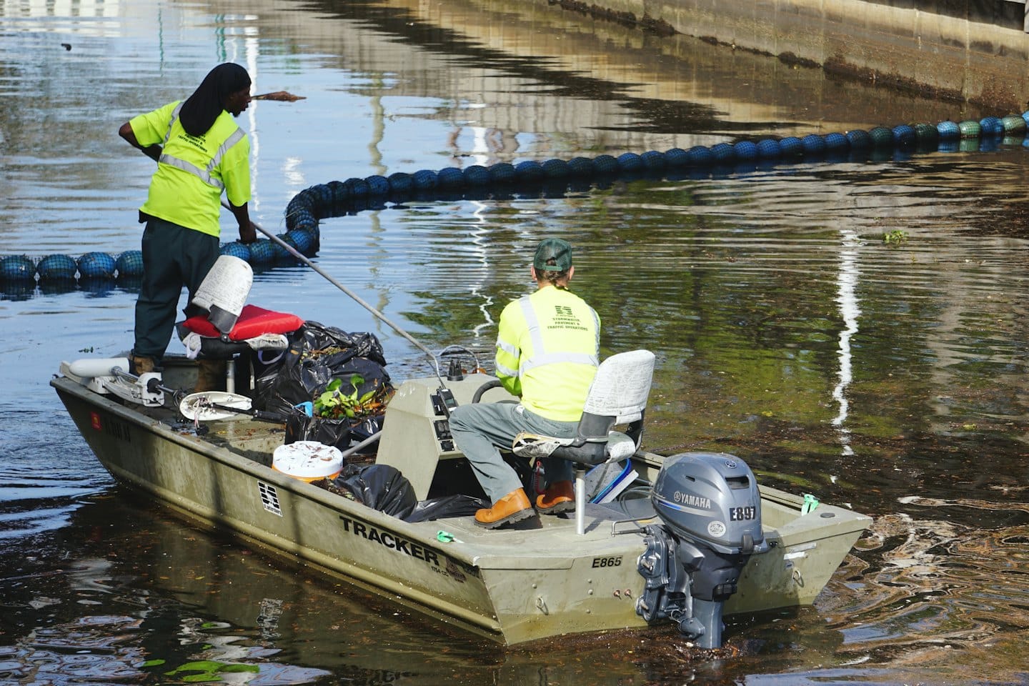 Environmental workers remove debris from a channel using a motorboat.