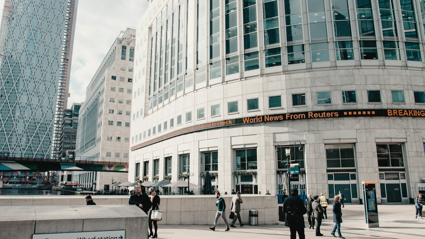 Pedestrians walk past modern city buildings with a news ticker.