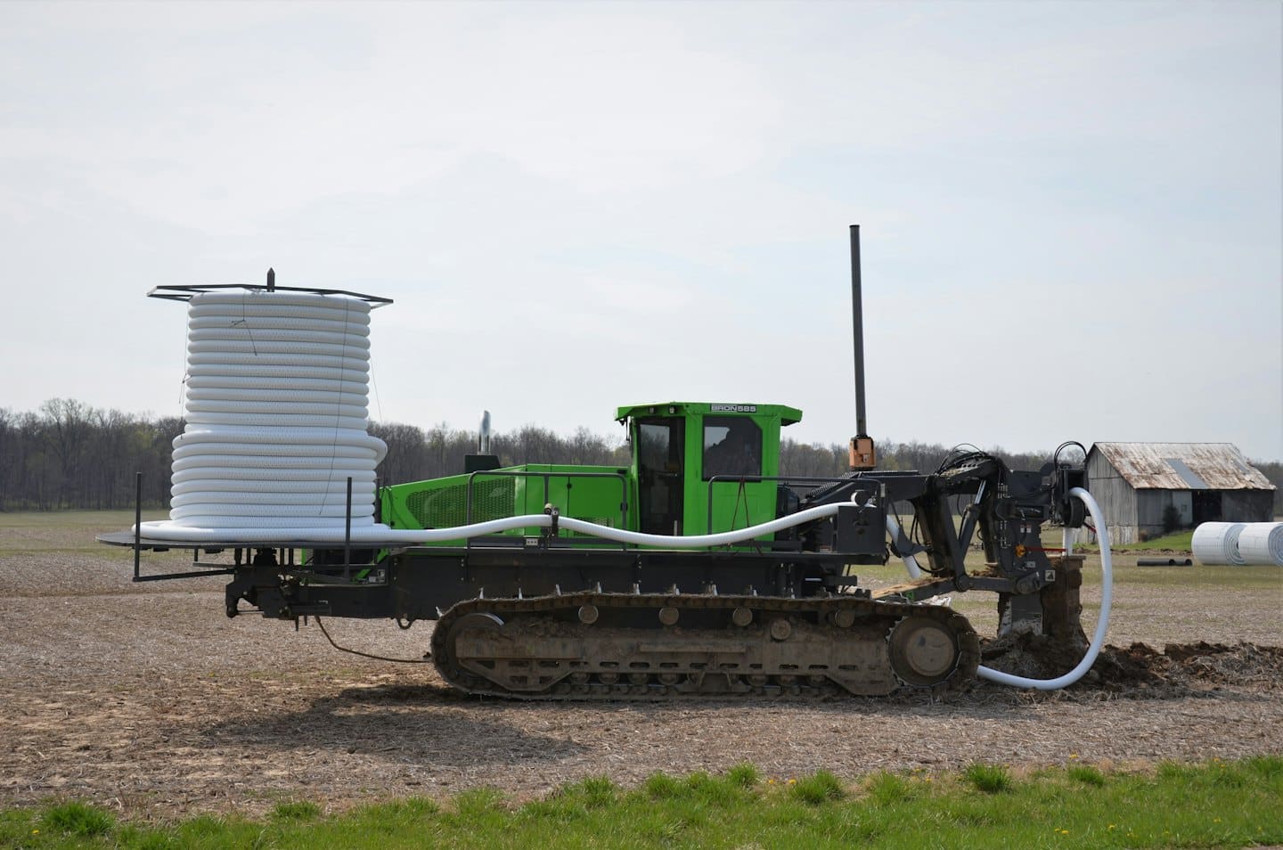 Green trenching machine laying white pipe in a brown field.