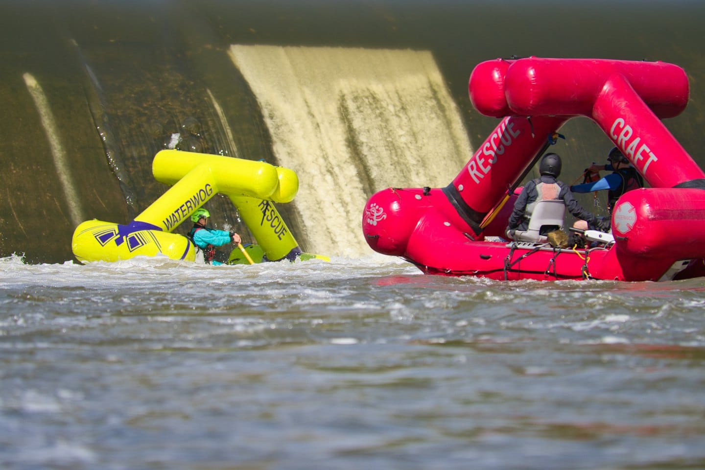 Three people in rescue rafts navigate turbulent water near a dam.